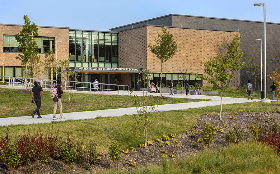 A large new brick building with students in uniform walking up a sidewalk towards the school entrance and a rain garden in the foreground.