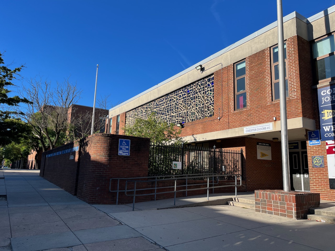 A modernist brick building with a large stained glass across the front, a flag pole next to an entrance on the right, and a low brick wall along the sidewalk on the left.